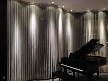 The stainless steel metal coil drapery is installed on the wall and a piano on the ground.